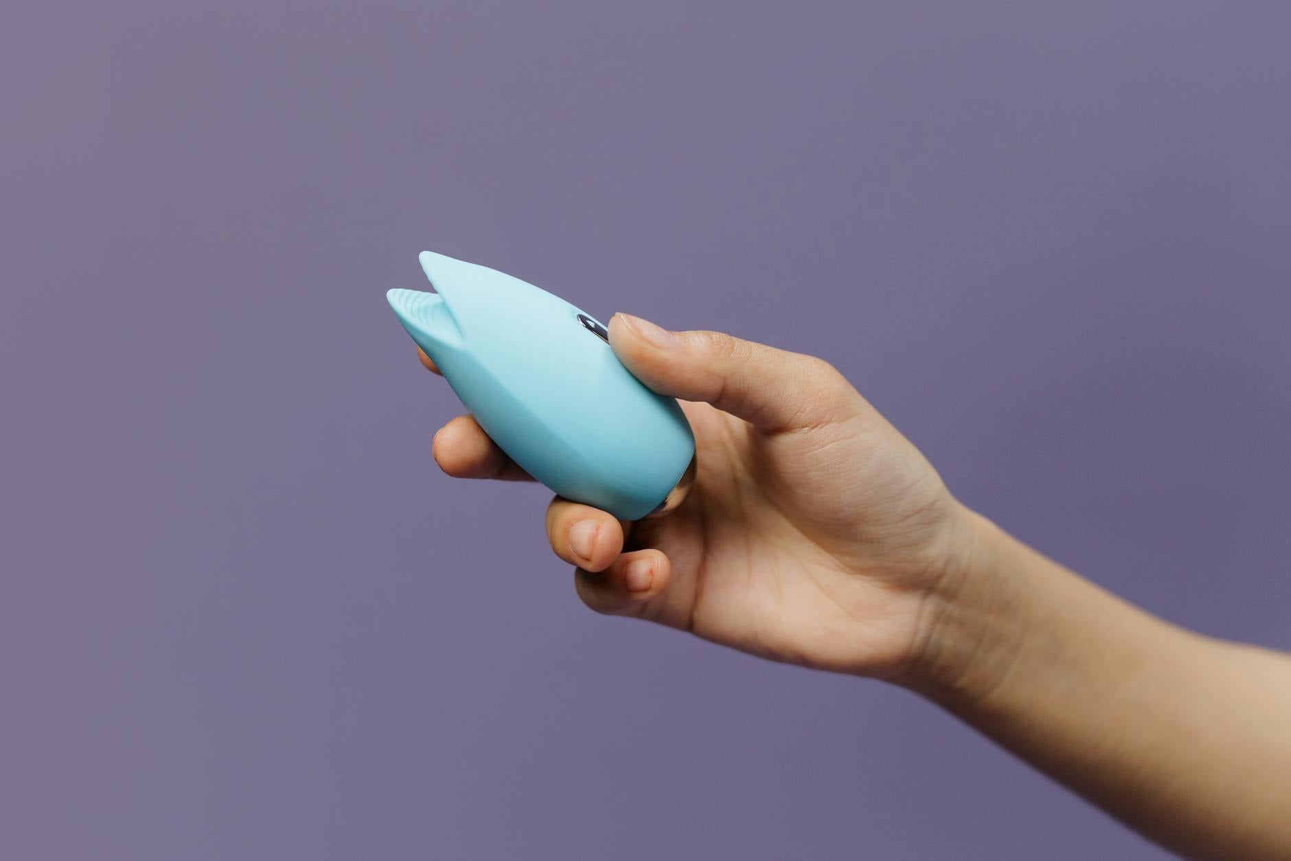 A silicone clitoral vibrator held in hand against a purple background, representing safe self-exploration
